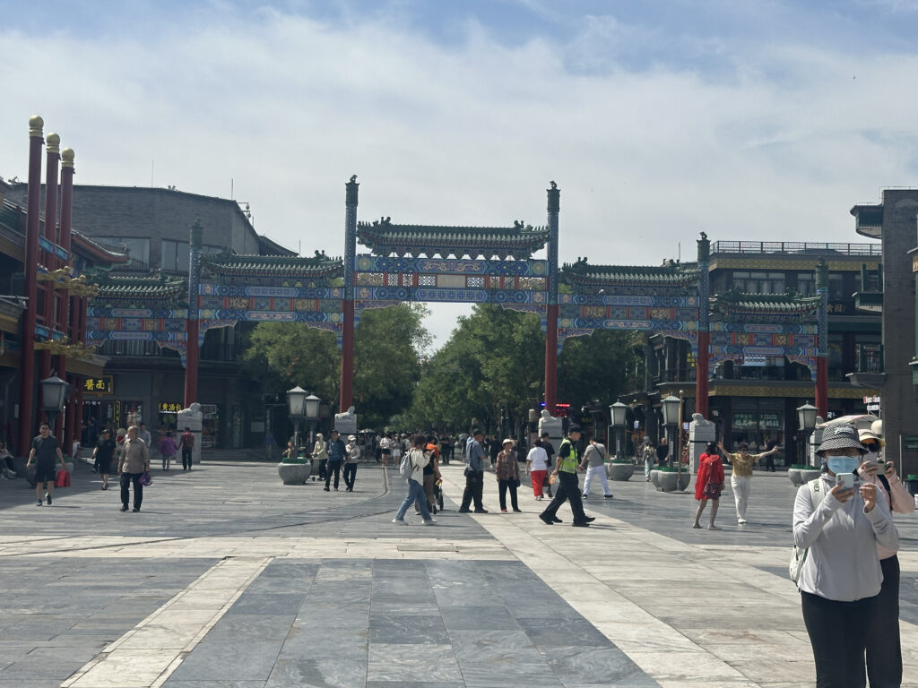 A bustling street scene featuring a traditional Chinese archway with intricate designs and vibrant colors. People are walking and taking photos, and there are buildings with traditional architectural elements on either side. The sky is partly cloudy, adding to the lively atmosphere.