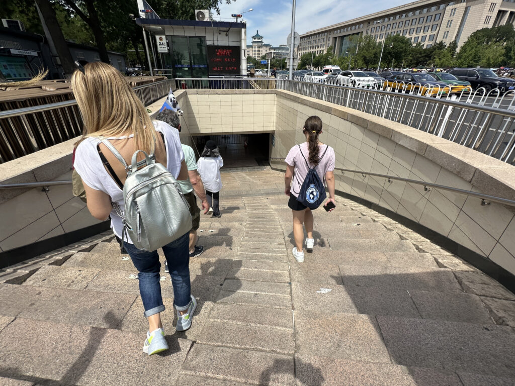 A group of people is walking down a set of stone stairs leading into an underground passage. The scene is outdoors, with a busy street and buildings visible in the background. The people are casually dressed, and some are carrying backpacks. The area is surrounded by railings and there is a sign with red text near the entrance.