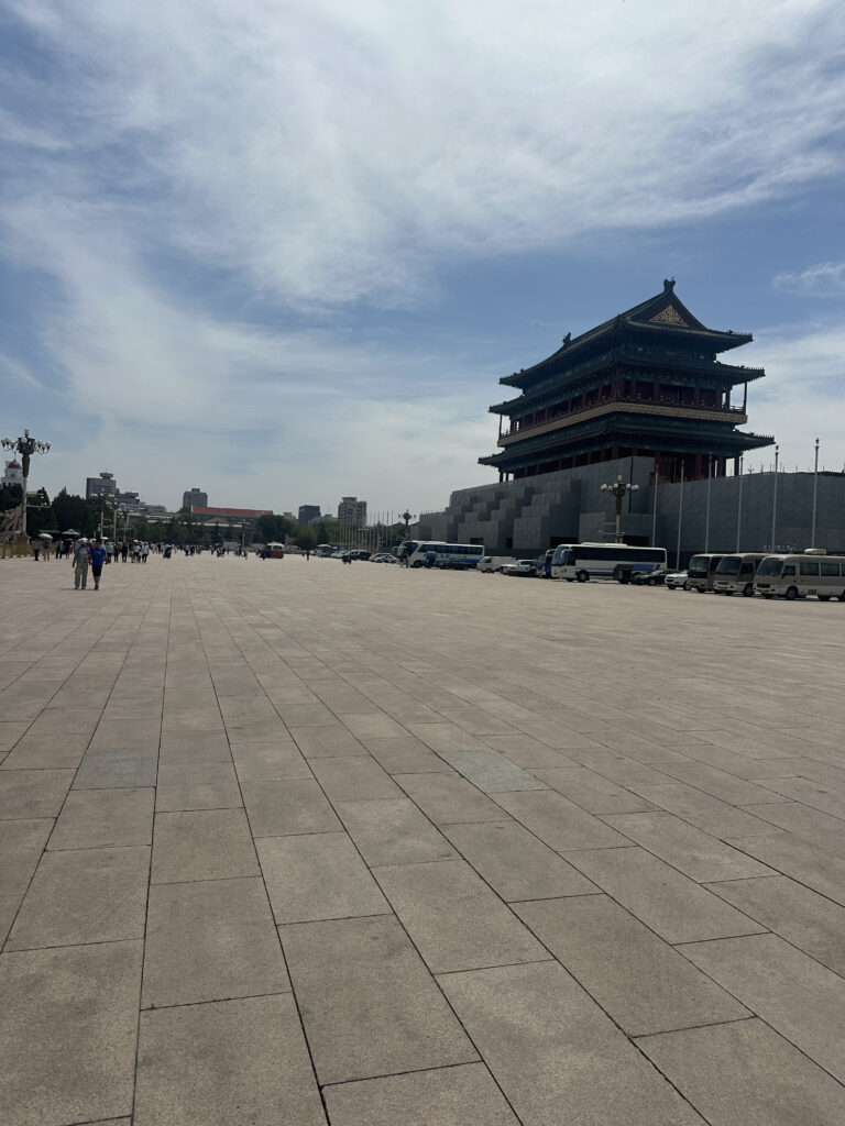 A large, open square with a traditional multi-tiered Chinese building on the right. The sky is partly cloudy, and there are a few people walking in the distance. Several vehicles are parked along the side of the square.