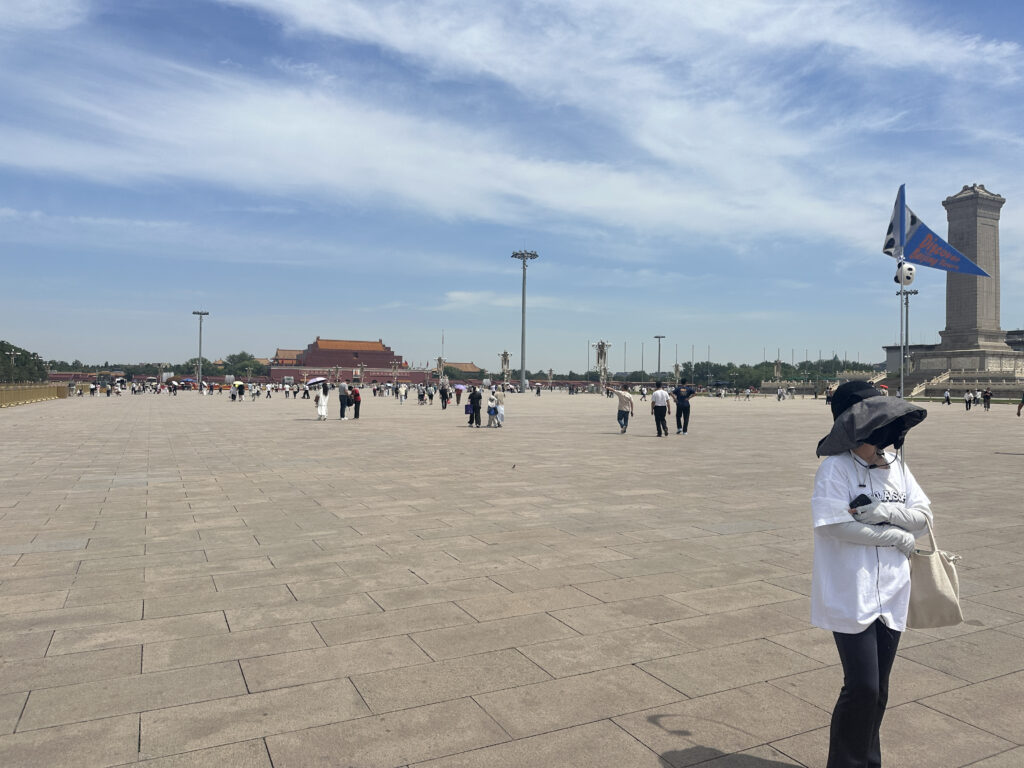 A large open square with a few people walking around. In the background, there is a traditional-style building with a red roof. A tall monument is visible on the right side. The sky is mostly clear with some clouds. In the foreground, a person wearing a wide-brimmed hat and a mask is standing, holding a bag.