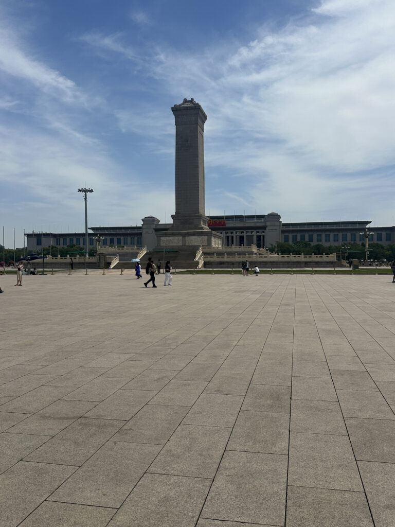 The image shows a large open square with a tall stone monument in the center. The monument is surrounded by a low fence, and there are a few people walking around the area. In the background, there is a large building with a flat roof. The sky is partly cloudy.