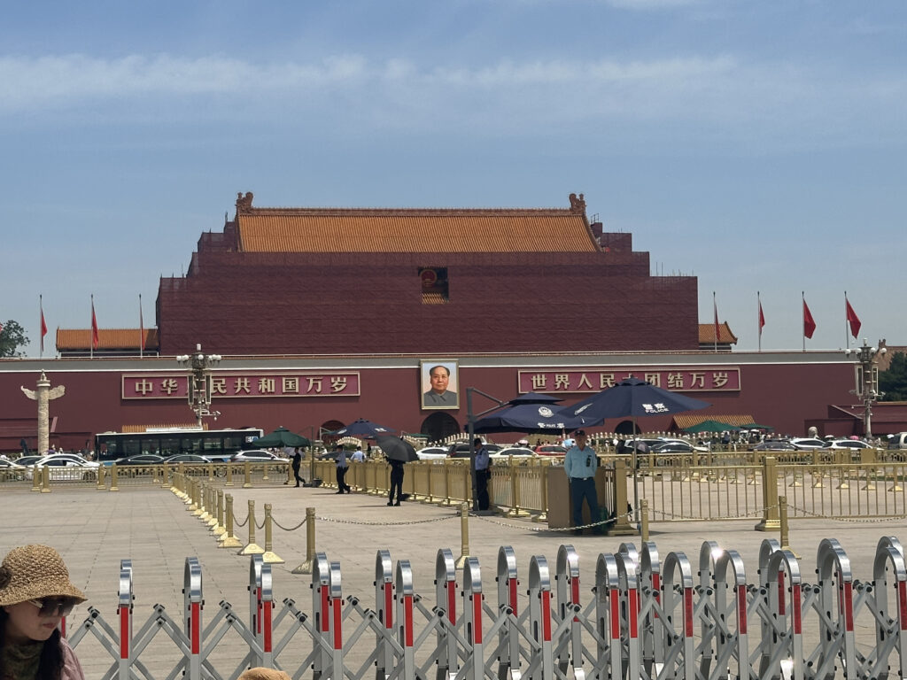 The image shows the Tiananmen Gate in Beijing, China. The gate features a large portrait of a man and is adorned with red flags. There are Chinese characters on the facade. In the foreground, there are barriers and people, some holding umbrellas, standing or walking around. The sky is clear and blue.
