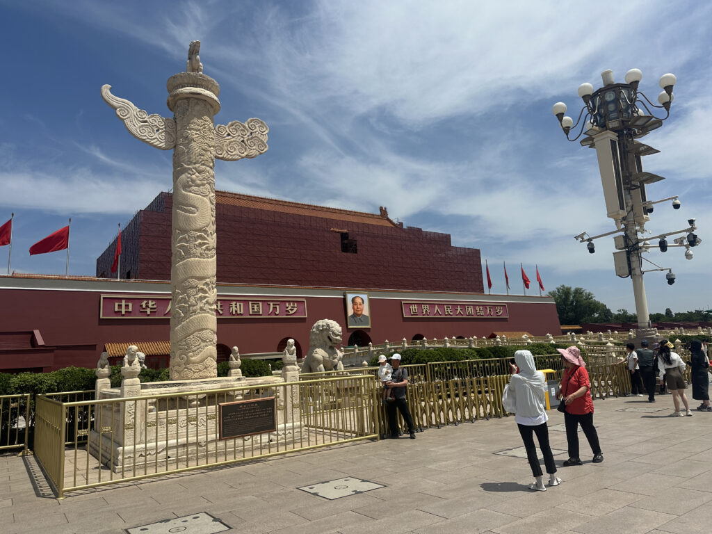 A large stone column with intricate carvings stands prominently in the foreground, surrounded by a metal fence. Behind it is a red building with a portrait and Chinese characters on the facade. Several red flags are visible on the building's roof. People are walking and standing around the area, and a tall streetlight with multiple lamps is on the right. The sky is clear with some clouds.