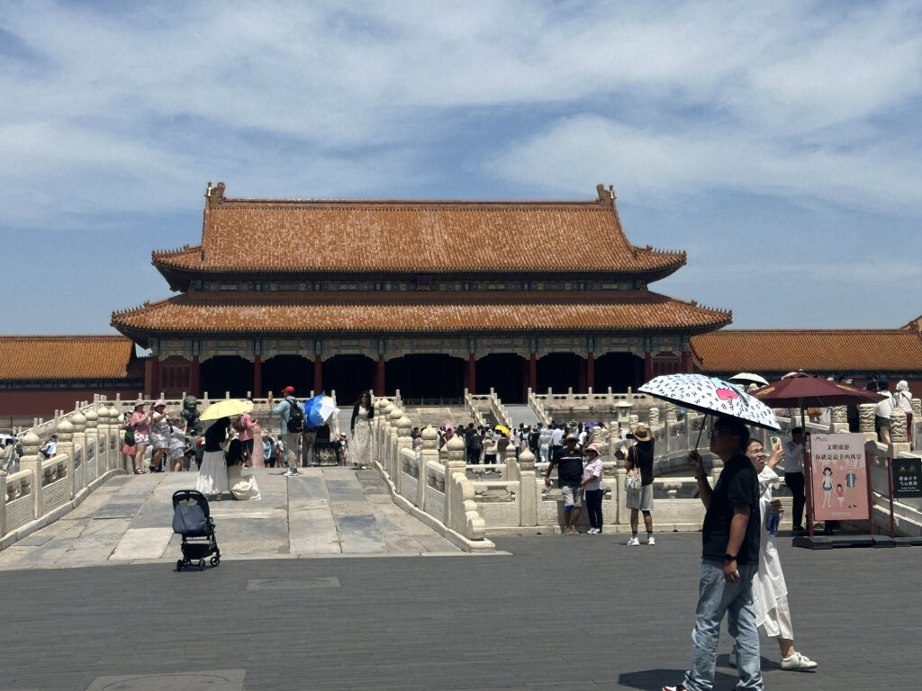 The image shows a large traditional Chinese building with a red and gold roof, likely part of the Forbidden City in Beijing. In the foreground, there are many people walking and standing on a stone bridge, some holding umbrellas for shade. The sky is clear with a few clouds.