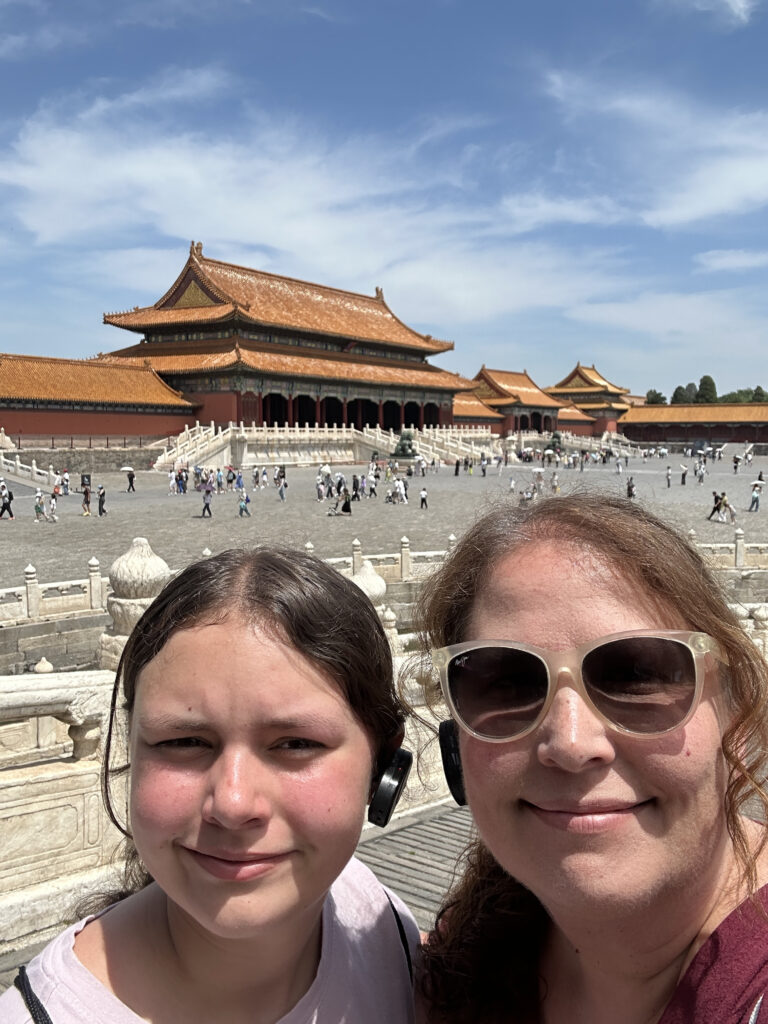 A selfie of two people in front of a large, traditional Chinese building with ornate, sloping roofs, likely part of the Forbidden City. The sky is clear and blue, and there are several people walking in the background. The person on the right is wearing sunglasses.