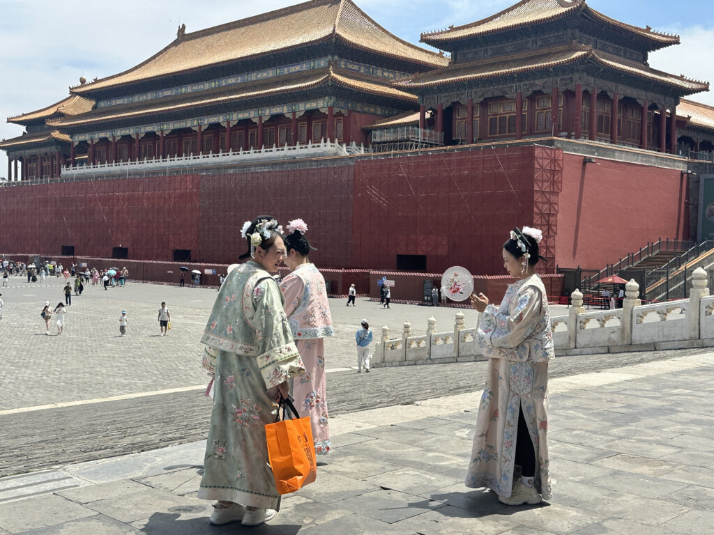 A group of people dressed in traditional Chinese clothing are standing in front of a large, historic building with ornate roofs, likely part of the Forbidden City in Beijing. The building features red walls and golden roofs. The individuals are wearing floral-patterned garments and hair accessories, and one person is holding a decorative fan. The scene is set on a sunny day with other visitors in the background.