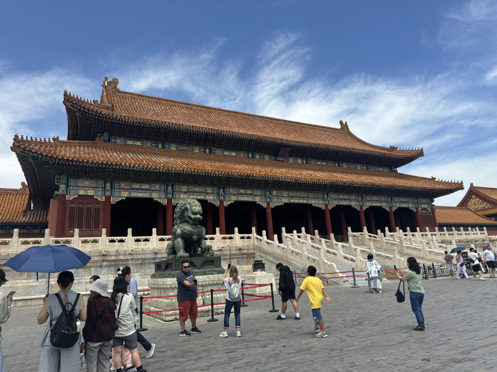 A large traditional Chinese building with ornate details and a sloping roof is in the background. In front of the building, there is a stone lion statue. Several people are walking and taking photos in the foreground. The sky is clear with a few clouds.