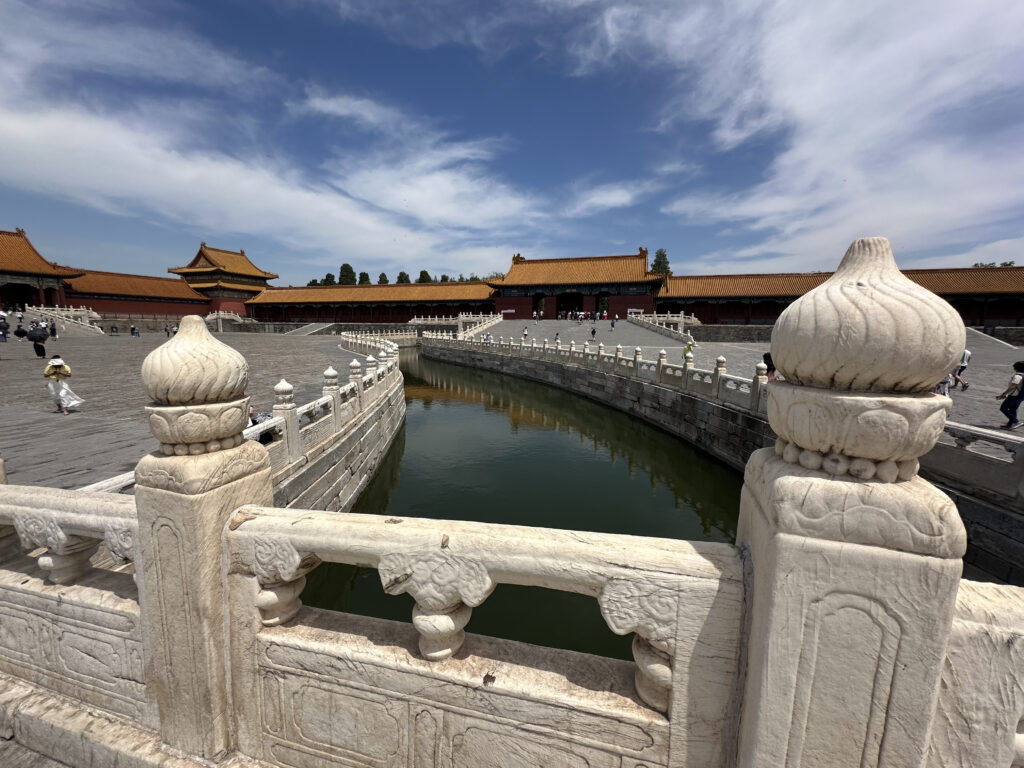The image shows a traditional Chinese architectural scene with a stone bridge featuring ornate carvings and decorative posts. The bridge spans over a narrow body of water. In the background, there are buildings with classic Chinese-style curved roofs and red walls. The sky is partly cloudy, and a few people are walking around the area.