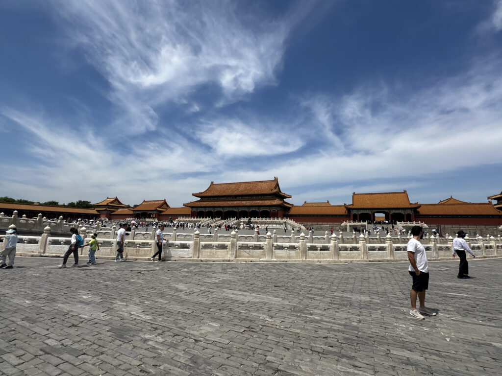 The image shows a large, open courtyard in front of traditional Chinese architecture with ornate, sloping roofs. The sky is clear with some clouds, and there are several people walking around the area, suggesting it is a popular tourist site. The buildings are surrounded by stone railings, and the ground is paved with large stone tiles.