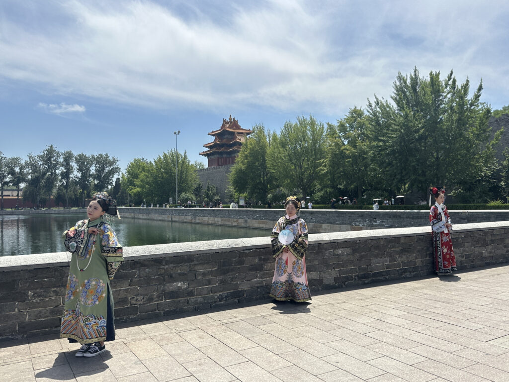 Three people are dressed in traditional Chinese clothing, standing along a stone wall by a body of water. The background features a traditional Chinese building with a multi-tiered roof and lush green trees under a partly cloudy sky.