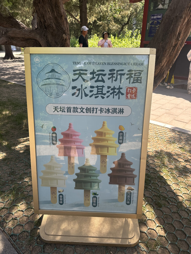 The image shows a signboard advertising "Temple of Heaven Blessing Ice Cream." The sign features illustrations of ice cream bars shaped like the Temple of Heaven, each in different colors and flavors. The flavors include strawberry, mango, vanilla, matcha, and chocolate. The sign is placed outdoors, with trees and people visible in the background.