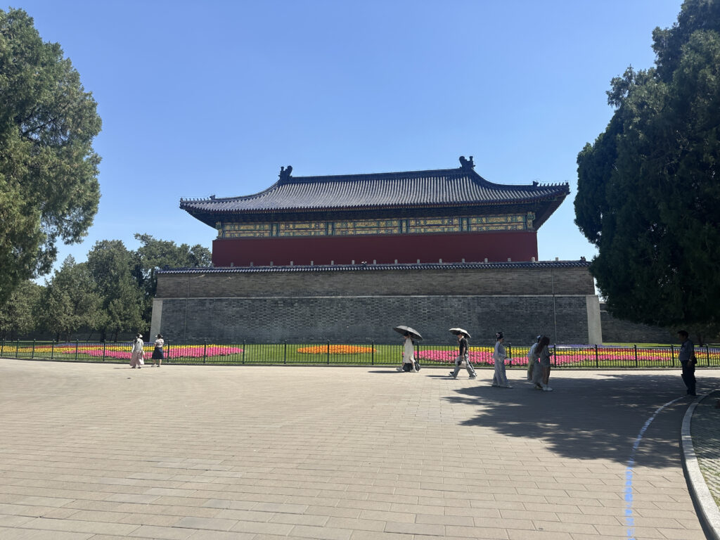 A traditional Chinese building with a curved roof and ornate details is in the center of the image. The building is surrounded by a stone wall and colorful flower beds. Several people are walking in front of the building, some holding umbrellas. Tall trees frame the scene on both sides, and the sky is clear and blue.