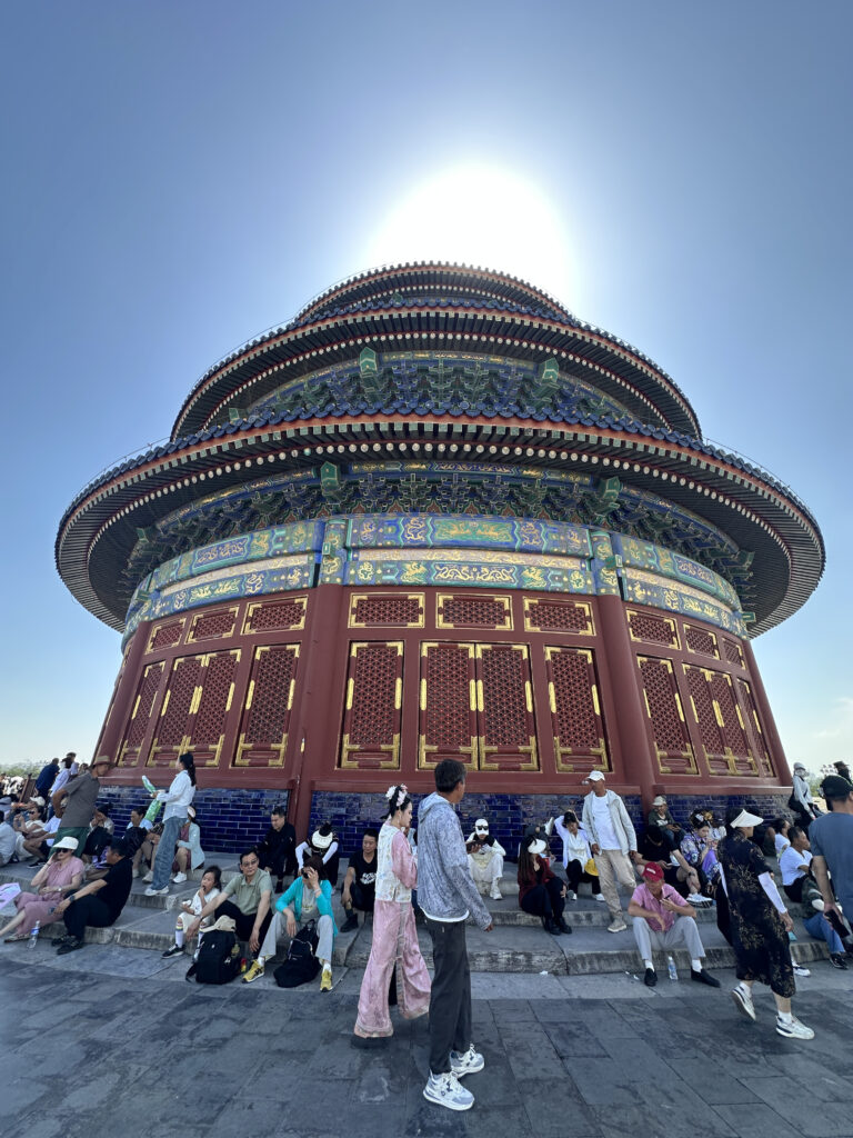 The image shows a large, ornate, circular building with intricate designs and vibrant colors, likely a traditional Chinese structure. The sun is positioned directly above, creating a halo effect. In the foreground, a group of people are gathered, some sitting on steps and others standing, with a few wearing traditional clothing. The sky is clear and blue.