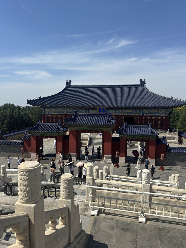 The image shows a traditional Chinese architectural structure with a large, ornate building featuring a blue-tiled roof and decorative details. In the foreground, there are stone railings and steps leading up to the building. Several people are walking around the area, some holding umbrellas. The sky is clear with a few clouds.