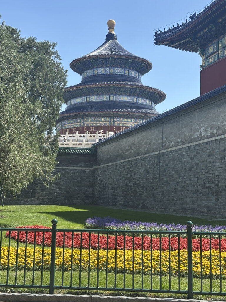 The image shows the Temple of Heaven, a historic circular building with a blue-tiled roof and intricate designs, located in Beijing, China. In the foreground, there is a well-maintained garden with colorful flowers arranged in rows of red, yellow, and purple. A green metal fence separates the garden from the pathway. A large tree is visible on the left side, and the sky is clear and blue.