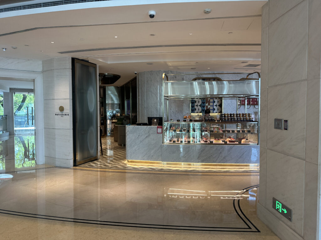 The image shows the entrance to a patisserie with a modern and elegant design. The interior features marble walls and floors, with a glass display case showcasing various pastries and desserts. There is a counter with a person behind it, and the area is well-lit with overhead lighting. The patisserie sign is visible on the left wall, and there is an emergency exit sign on the right.