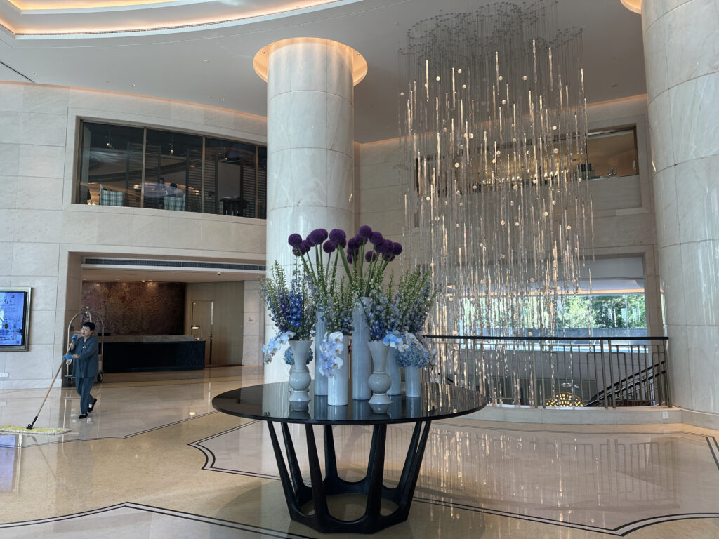 The image shows a luxurious hotel lobby with a large, elegant chandelier hanging from the ceiling. In the foreground, there is a round black table with several vases containing purple and blue flowers. The lobby features marble columns and walls, and there is a person cleaning the floor with a mop. A reception desk is visible in the background, along with a luggage cart. The space is well-lit with natural light coming through large windows.