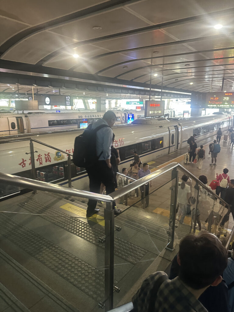 A busy train station with people walking down a staircase towards a platform. A train is visible on the tracks, and there are signs and lights overhead. The station has a modern design with a curved ceiling.