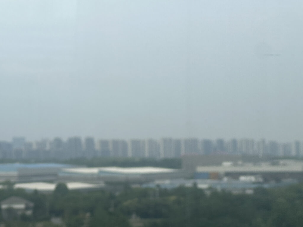 A hazy cityscape with a row of tall buildings in the background. In the foreground, there are several large, flat-roofed industrial or commercial buildings surrounded by greenery. The sky is overcast, contributing to the overall hazy appearance.