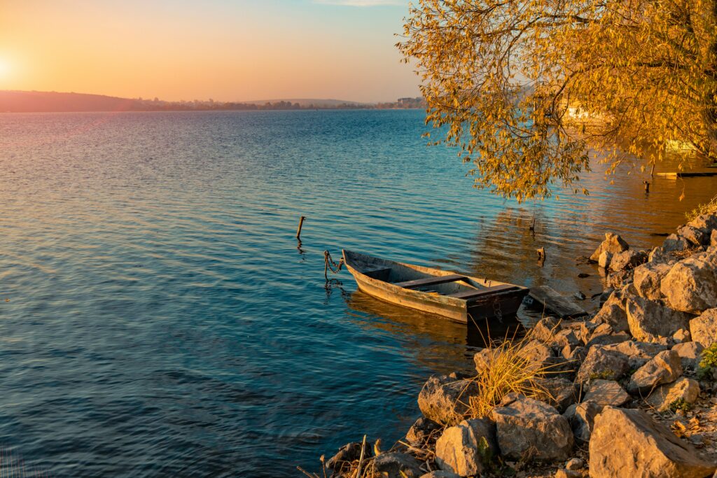 A small wooden boat is moored near the rocky shore of a calm lake during sunset. The water reflects the warm colors of the sky, and a tree with golden leaves overhangs the scene. The distant shoreline is visible under the soft, orange glow of the setting sun.