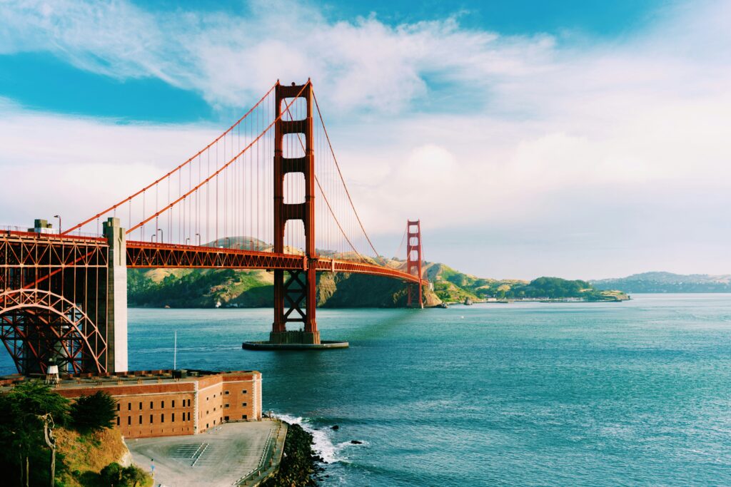 A scenic view of the Golden Gate Bridge in San Francisco, California. The iconic red suspension bridge spans across the blue waters of the bay, with green hills and a partly cloudy sky in the background. A fort-like structure is visible in the foreground near the water's edge.