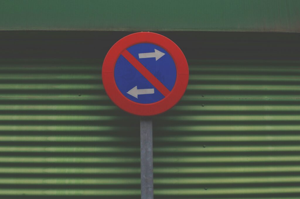 A circular traffic sign with a red border and a blue center, featuring a red diagonal line crossing through two white arrows pointing left and right, indicating no left or right turns. The sign is mounted on a metal pole against a green corrugated background.
