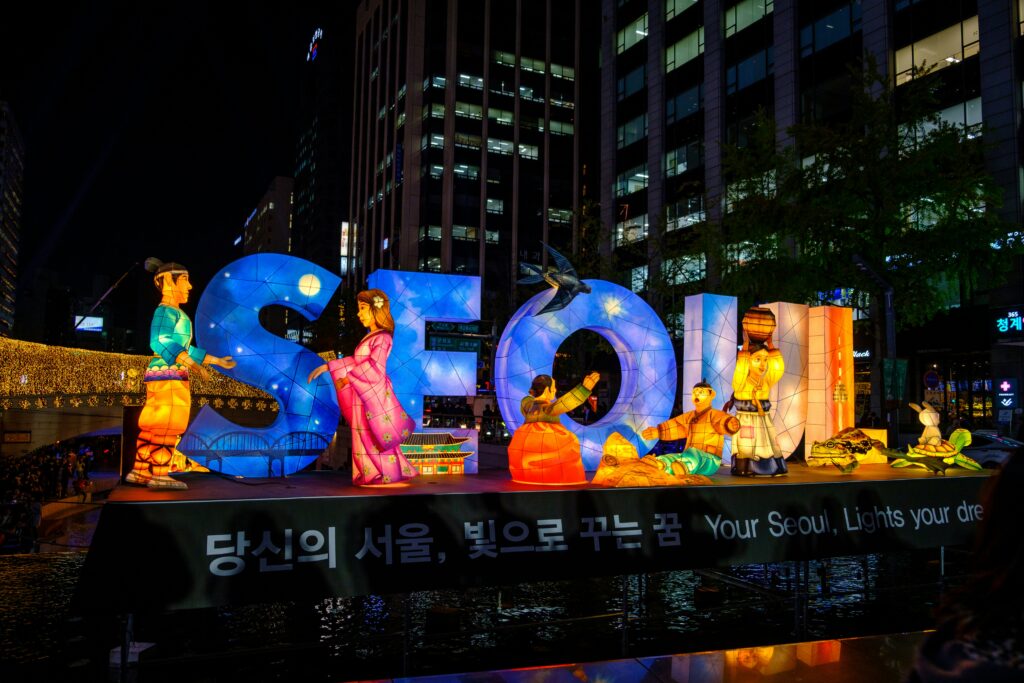The image shows a vibrant nighttime scene featuring large illuminated letters spelling "SEOUL." The letters are decorated with colorful lanterns depicting traditional Korean figures in various poses, including a man and woman in traditional attire, and other figures engaged in cultural activities. The background includes tall buildings with lit windows, and there is a banner at the bottom with text in both Korean and English, reading "Your Seoul, Lights your dream."