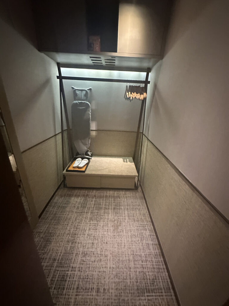 The image shows a small, narrow closet space with a carpeted floor. On the left side, there is an ironing board with an iron on top. A horizontal rod is mounted above, holding several wooden hangers. Below the ironing board, there is a bench with a pair of white slippers placed on it. The walls are plain, and there is a shelf above the rod. The lighting is soft and ambient.