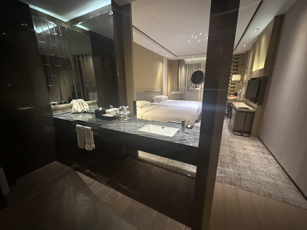 The image shows a modern hotel room with a view from the bathroom area. The bathroom features a large mirror and a countertop with a sink, towels, and bottled water. Beyond the bathroom, there is a bedroom with a neatly made bed, a round mirror, and a television mounted on the wall. The room has a contemporary design with soft lighting and neutral colors.