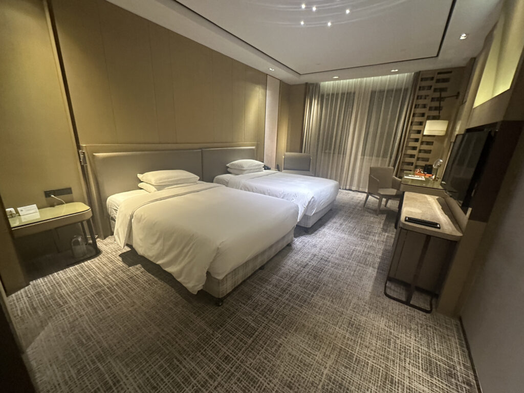 The image shows a modern hotel room with two neatly made single beds. The room features a neutral color palette with beige walls and carpet. There is a nightstand between the beds, a desk with a chair near the window, and a flat-screen TV mounted on the wall. The window is covered with sheer curtains, and the room is well-lit with ceiling lights.