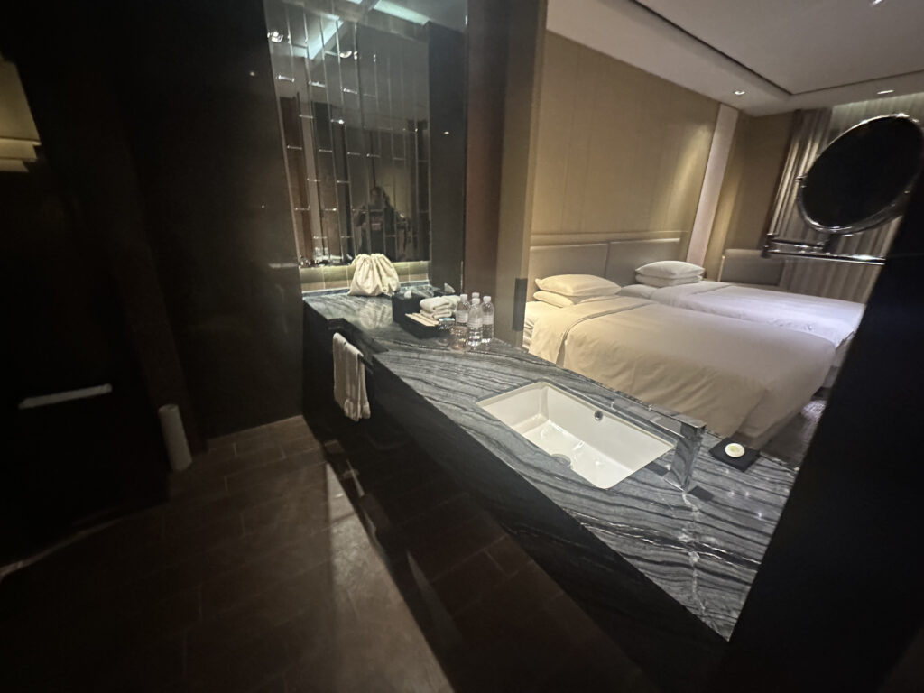 The image shows a hotel room with a modern design. In the foreground, there is a marble countertop with a built-in sink, towels, and bottled water. A large mirror is mounted above the countertop. In the background, there are two neatly made beds with white linens. The room has soft lighting and a neutral color palette, creating a cozy and elegant atmosphere.