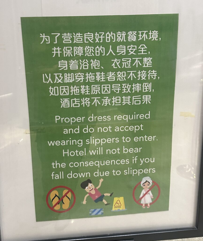 The image shows a sign with text in both Chinese and English. The sign states that proper dress is required and slippers are not allowed. It also mentions that the hotel will not be responsible for any consequences if someone falls due to wearing slippers. The sign includes illustrations: a person slipping, a pair of slippers with a prohibition symbol, and a person in a bathrobe with a prohibition symbol. There is also a caution sign depicted. The background of the sign is green.