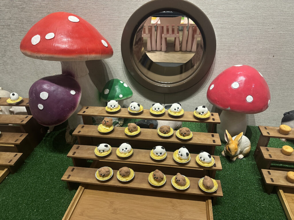 The image shows a whimsical display featuring large, colorful mushroom sculptures in red, purple, and green with white spots. In front of the mushrooms, there are wooden shelves holding small, round pastries decorated to look like cute animal faces, including pandas and other creatures. A small rabbit figurine is also part of the display, which is set on a green, grass-like surface. A circular mirror is mounted on the wall behind the setup.