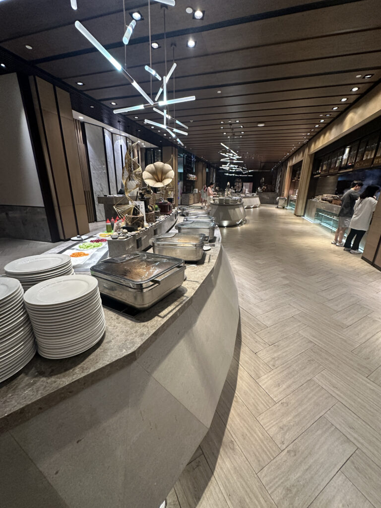 The image shows a modern buffet setup in a restaurant. There are stacks of white plates on a counter, with several chafing dishes containing food. Decorative elements, including a sculpture and a gramophone-like object, are placed on the counter. The ceiling features contemporary lighting fixtures, and the floor has a herringbone pattern. In the background, a few people are seen near the buffet area.