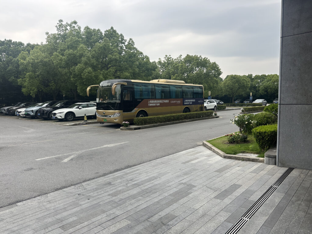 A parking lot with several cars parked in a row next to a large bus. The bus is gold and blue with some text on its side. There are trees in the background and a sidewalk with neatly trimmed bushes and flowers in the foreground. The sky is overcast.