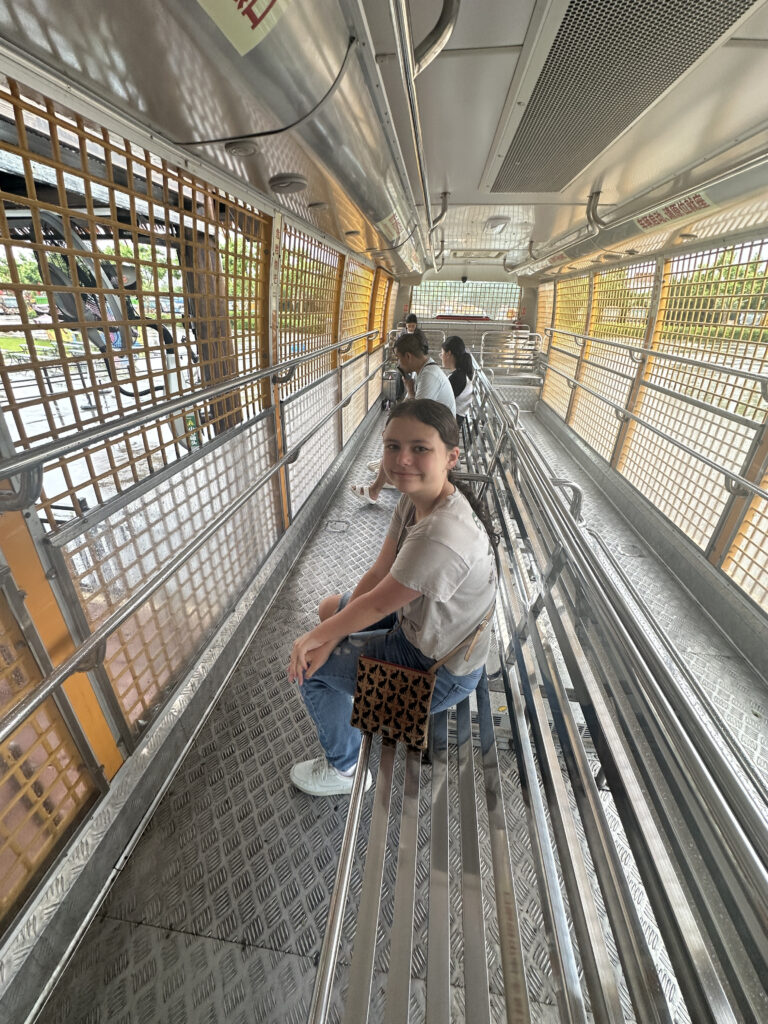 A person is sitting inside a metal tram or transport vehicle with grated windows and metal benches. The interior is spacious, with several people seated along the sides. The person in the foreground is wearing a cap, a light-colored shirt, jeans, and white sneakers, and is carrying a patterned bag. The vehicle appears to be stationary, with a view of the outside through the windows.
