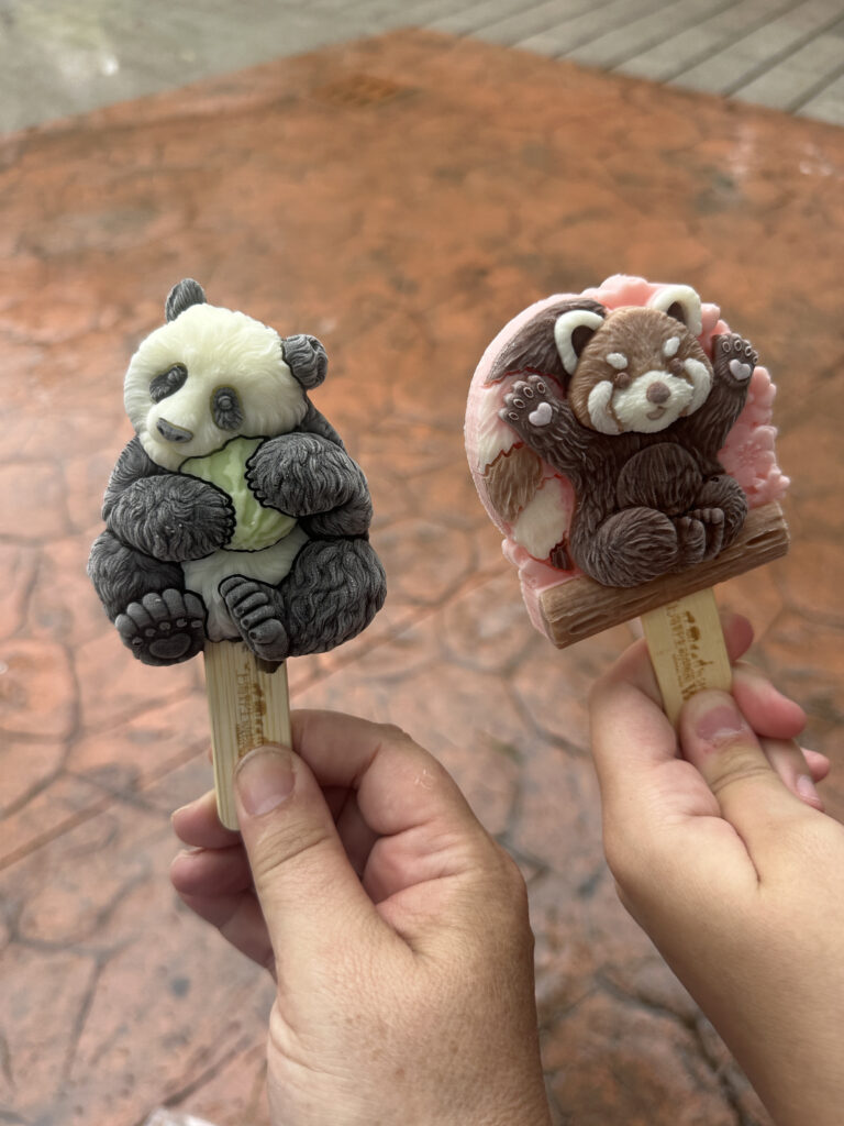 The image shows two intricately designed ice cream bars shaped like animals. The ice cream on the left is shaped like a panda holding a piece of bamboo, with detailed features in black and white. The ice cream on the right resembles a red panda, with a mix of brown, white, and pink colors, and is posed playfully. Both ice creams are held by wooden sticks, and two hands are visible holding them against a textured, reddish-brown background.