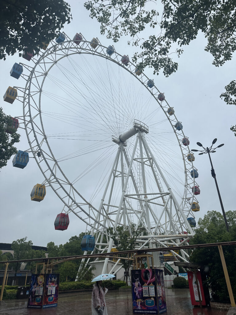 A large Ferris wheel with colorful gondolas is set against a cloudy sky. The structure is surrounded by trees, and a person holding an umbrella is walking in the foreground. There are posters and signs near the base of the Ferris wheel.