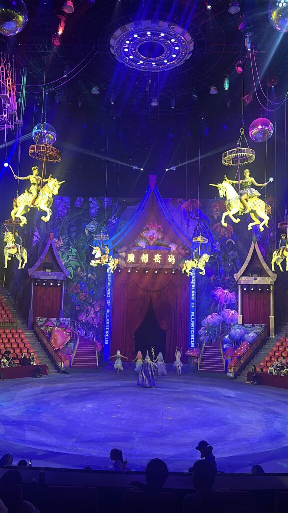 The image shows a vibrant circus stage with a colorful, fantasy-themed backdrop. Several performers are suspended in the air on glowing carousel horses, while others are on the stage below, dressed in elaborate costumes. The stage is surrounded by red seating, and the ceiling is adorned with bright lights and decorations. The atmosphere is lively and magical, with a large red curtain and whimsical elements enhancing the scene.