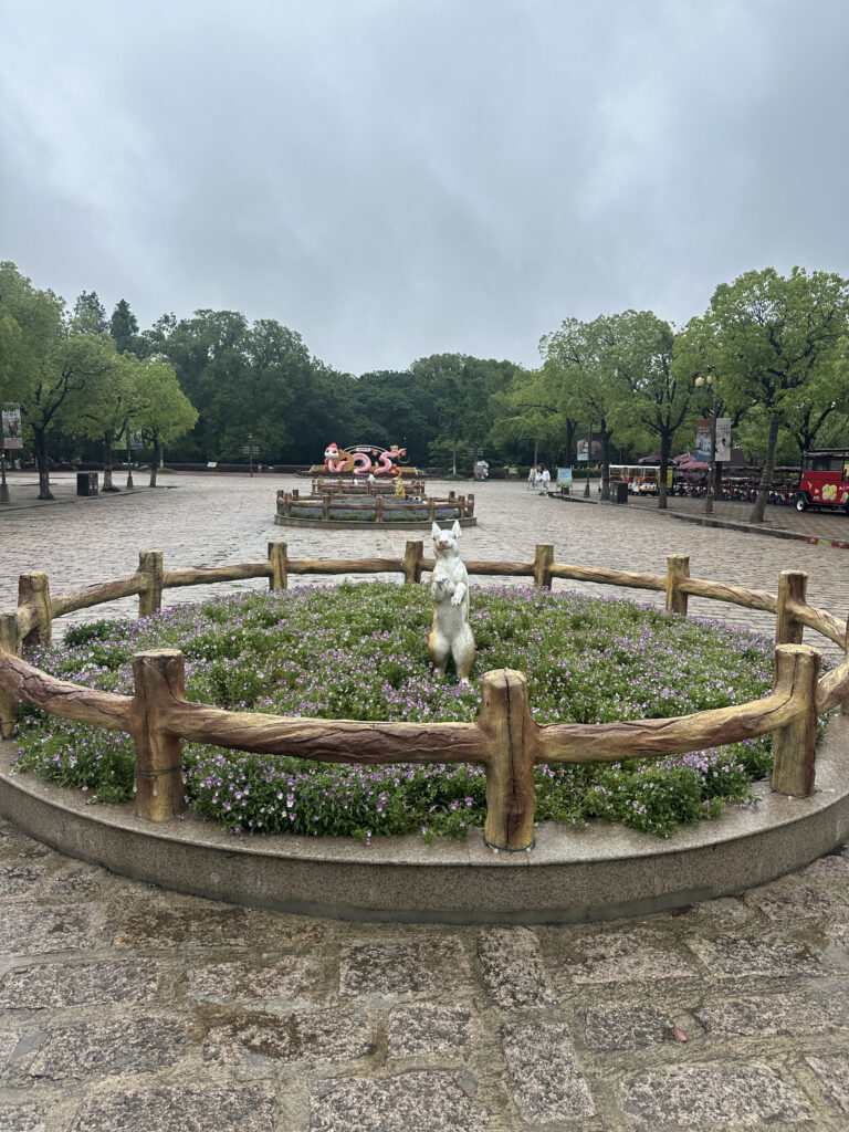 A circular flower bed with a wooden fence surrounds a small statue of a standing animal, possibly a squirrel, in the center. The flower bed is filled with green plants and small purple flowers. In the background, there is a larger sculpture of a pink octopus with a hat and sunglasses. The area is paved with stone tiles, and there are trees and a cloudy sky in the background. A red vehicle is parked on the right side.