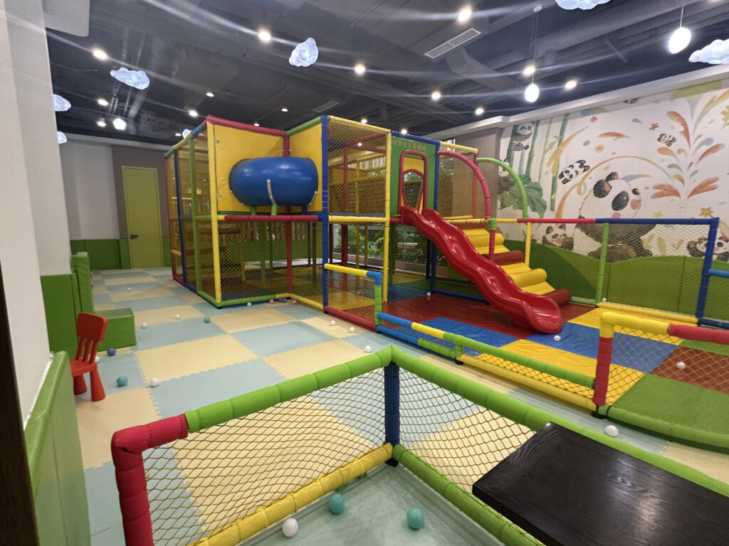 The image shows an indoor children's play area with colorful padded flooring and structures. There is a red slide attached to a multi-level play structure with yellow, blue, and red elements. The area is enclosed with netting for safety. The walls are decorated with playful murals featuring pandas and nature themes. The ceiling has cloud-shaped decorations and hanging lights. There are scattered plastic balls on the floor, and a small red chair is visible in the corner.