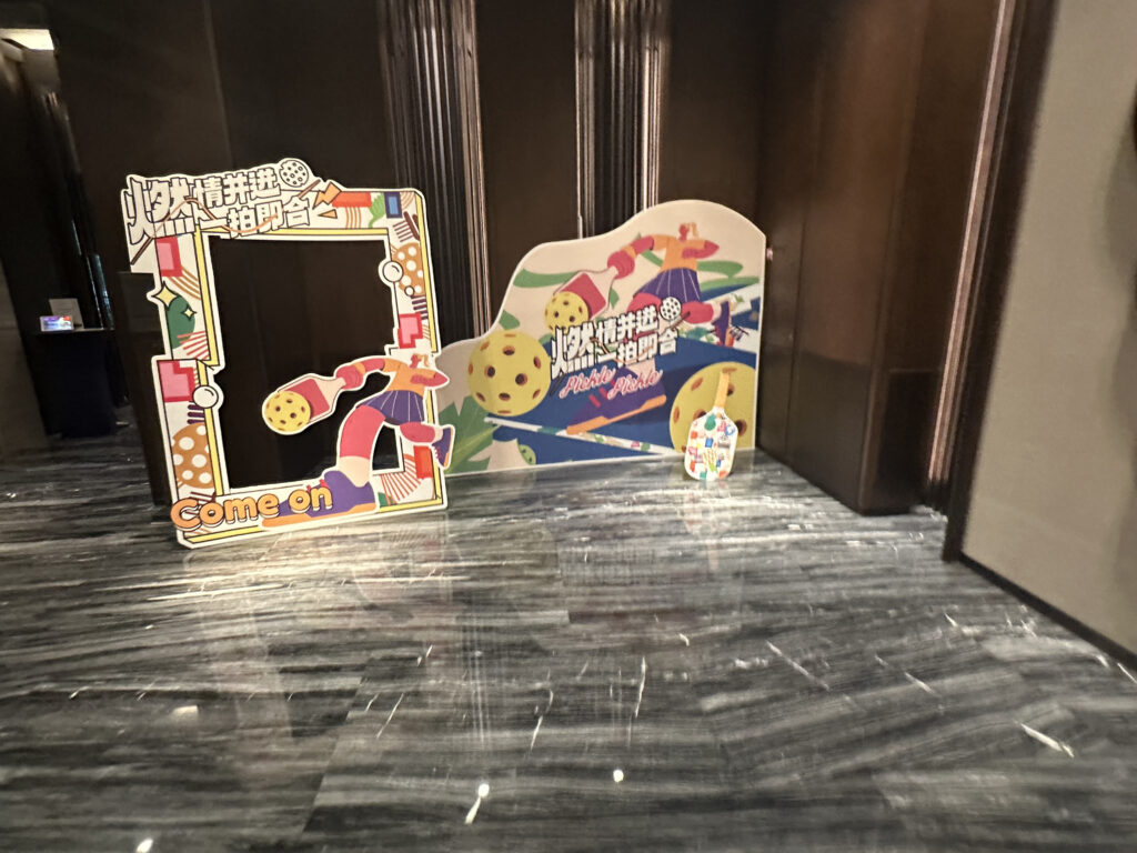 The image shows a colorful photo booth setup with two large cutouts. The left cutout is a rectangular frame with playful designs and the words "Come on" at the bottom. The right cutout features abstract shapes and figures, with text in a different language. The floor is shiny and reflective, and the background consists of dark walls.