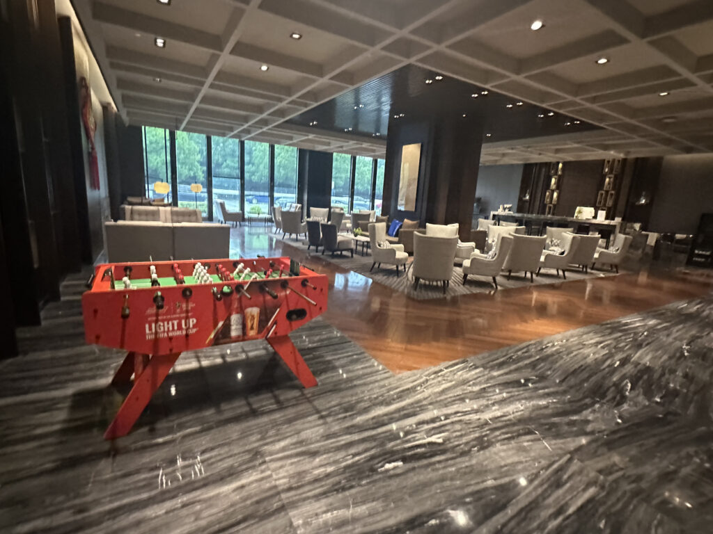 The image shows a spacious, modern lounge area with a foosball table in the foreground. The foosball table is red and has a sign related to the FIFA World Cup. In the background, there are several seating areas with armchairs and tables, set on a wooden floor. Large windows allow natural light to enter, and the ceiling has a grid pattern with recessed lighting.