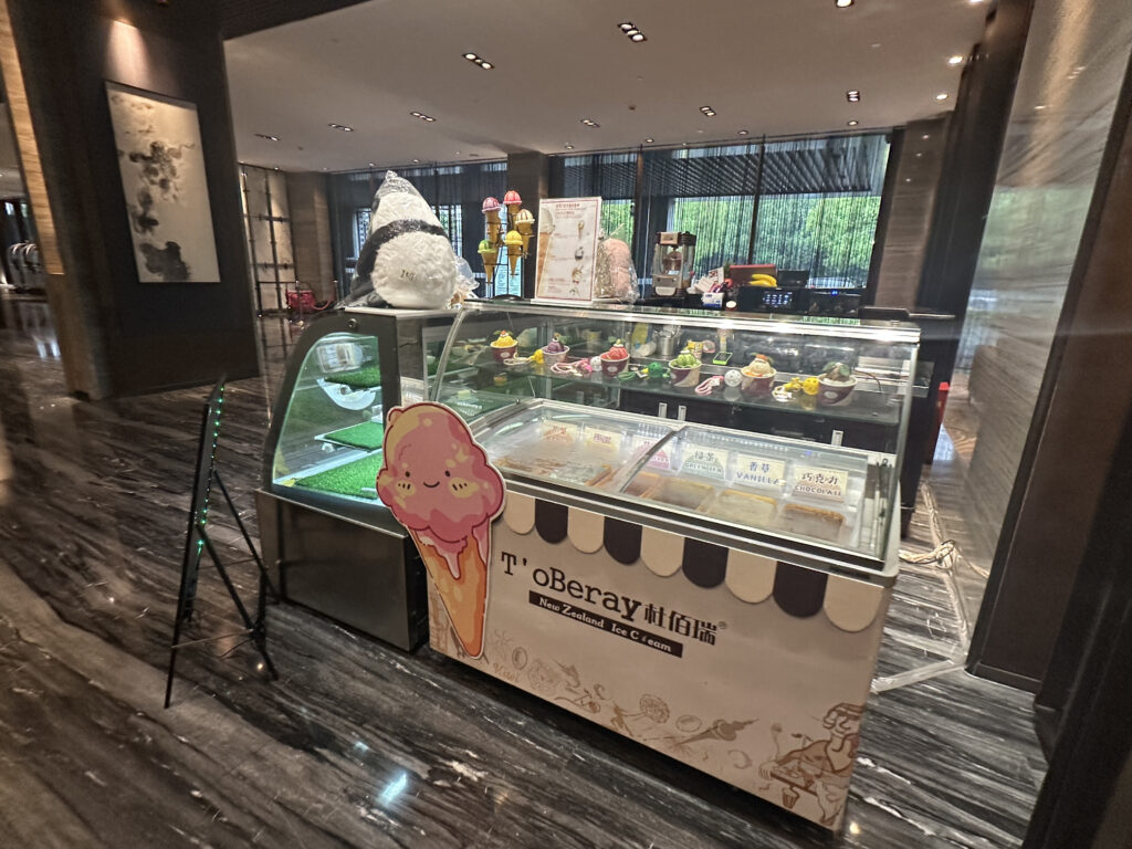 The image shows an ice cream display counter in a modern indoor setting. The counter has a glass top showcasing various ice cream flavors in cups, with colorful toppings. There is a large decorative ice cream cone figure on top of the counter, and a cute cartoon ice cream cone illustration on the front. The counter is labeled "T' oBeray New Zealand Ice Cream." The surrounding area has a polished marble floor and contemporary decor.