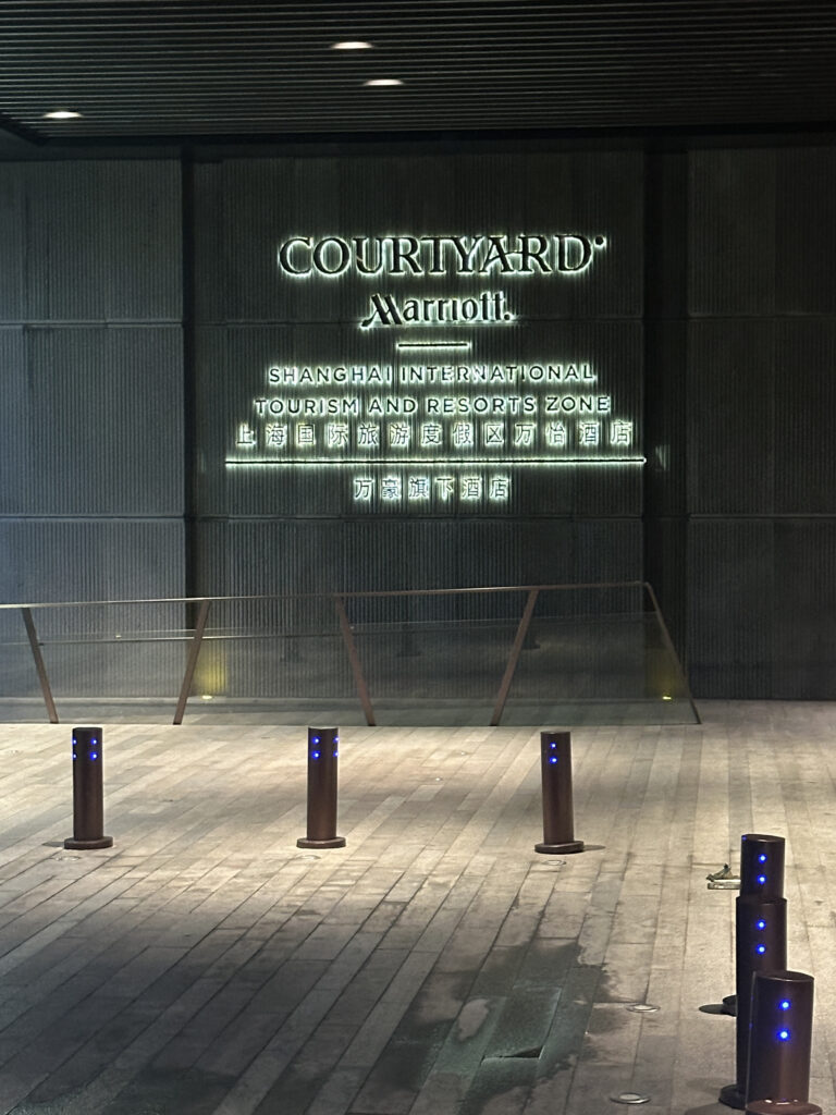 The image shows the entrance area of a building with a sign that reads "Courtyard Marriott" and "Shanghai International Tourism and Resorts Zone" in both English and Chinese. The area is well-lit, with several short bollards with blue lights lining the pathway. The ground is paved with large tiles, and there is a glass railing in the background.