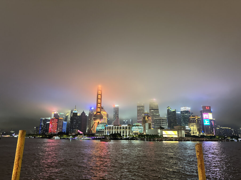 A nighttime cityscape featuring a brightly lit skyline with modern skyscrapers. The buildings are illuminated with colorful lights, and a prominent tower with a spherical structure is visible. The sky is overcast, and the city lights reflect on the water in the foreground.