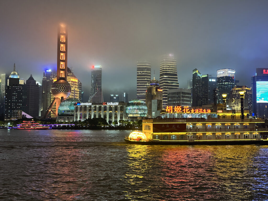 The image shows a nighttime cityscape of Shanghai, featuring the illuminated skyline of the Pudong district. Prominent is the Oriental Pearl Tower with its distinctive spherical design, surrounded by other modern skyscrapers. In the foreground, a brightly lit boat is sailing on the Huangpu River, reflecting colorful lights on the water. The sky is overcast, adding a misty ambiance to the scene.