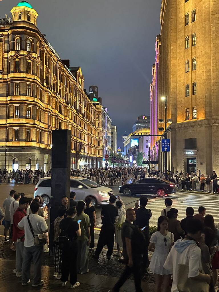 A bustling city street at night, with a large crowd of people gathered on the sidewalks. The street is lined with illuminated historic buildings, and cars are visible on the road. The scene is lively, with people taking photos and looking around. The buildings are lit with warm lights, creating a vibrant atmosphere.