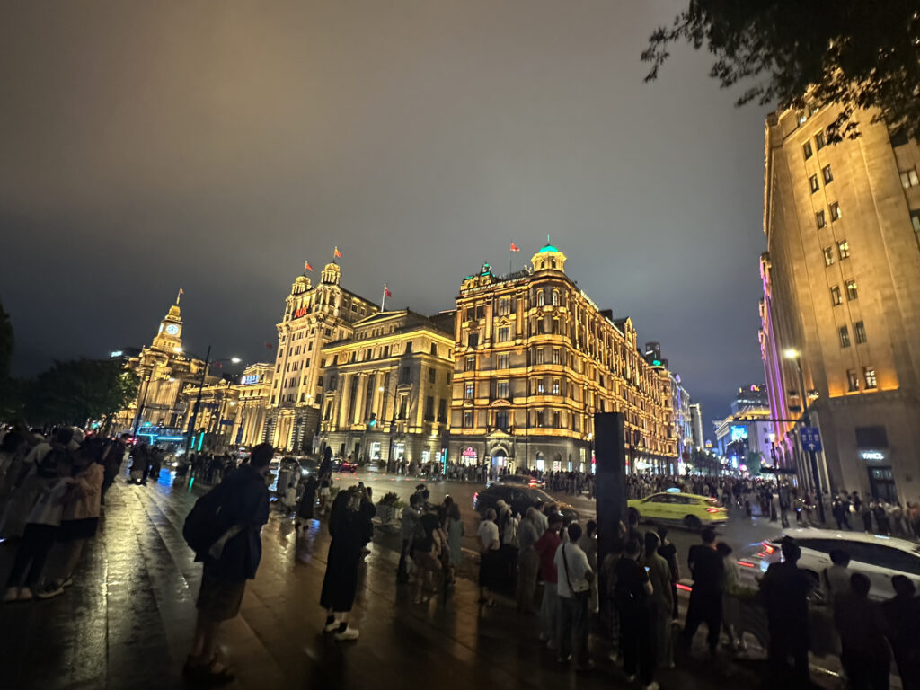 A bustling nighttime city scene with a large crowd of people gathered on a wet street. The buildings are illuminated with warm lights, showcasing intricate architectural details. The sky is overcast, and several cars, including a yellow taxi, are visible on the road. The atmosphere is lively and vibrant.
