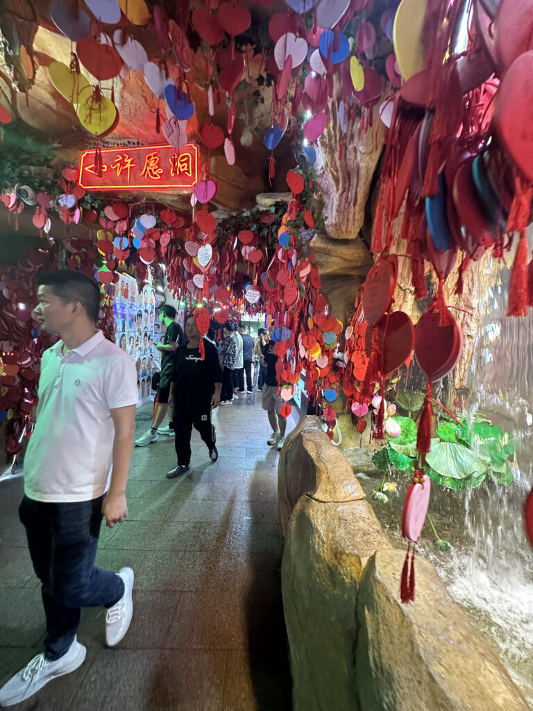 The image shows a narrow passageway decorated with numerous colorful hanging ornaments, primarily red, shaped like hearts and circles. A neon sign with Chinese characters is visible at the top. People are walking through the passage, and there is a stone wall with water and green plants on the right side. The atmosphere appears vibrant and festive.